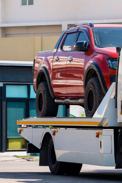 Broken red pickup car loaded onto white towing truck in public area for transportation to garage